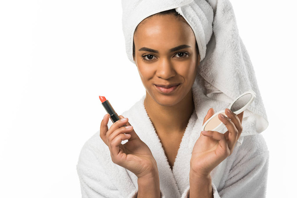 hermosa mujer afroamericana en albornoz y toalla sosteniendo lápiz labial y espejo, aislado en blanco
 - Foto, Imagen