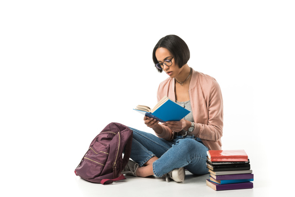 female african american student reading books while sitting on floor with backpack, isolated on white  - Photo, Image