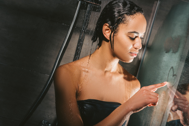 attractive african american woman taking shower and drawing heart sign on glass - Foto, imagen