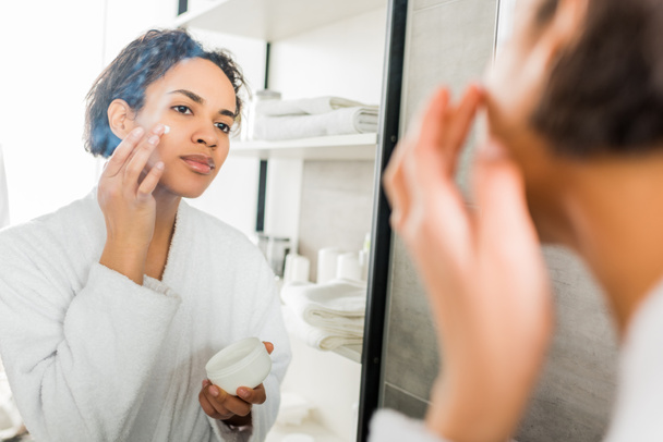 enfoque selectivo de chica afroamericana aplicando crema hidratante en la cara en el baño cerca del espejo
 - Foto, imagen