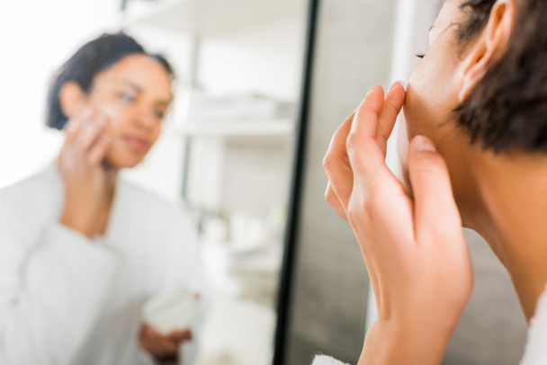 enfoque selectivo de la mujer afroamericana aplicando crema cosmética en la cara en el baño
 - Foto, Imagen
