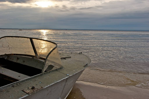Uma bela foto de um velho barco em uma praia tranquila. O sol nascente é visível através das nuvens e deixa uma bela reflexão sobre a água parada. Sereno
. - Foto, Imagem