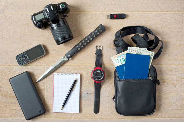 Ready-to-go spy kit.Photo camera, phone, powerbank, notepad with pen, knife, hand watch, USB memory stick, document bag with two passports and money in different currencies.Light wooden background. - Photo, Image