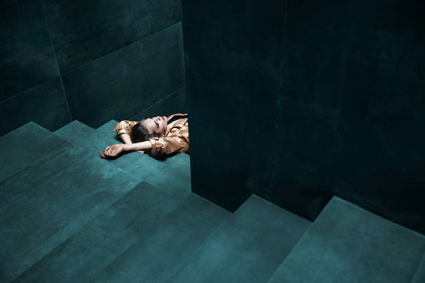 Amazing young woman in gold trench coat, black bodysuit with gold belt and in black polka dot tights is lying on the turquoise stairs - Foto, Imagen