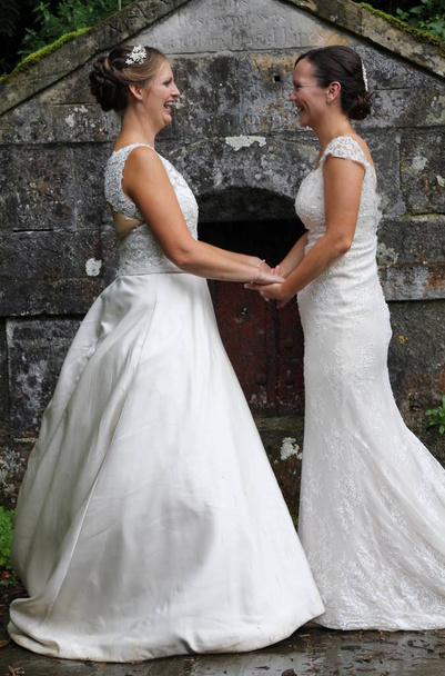 Two beautiful brides on their same sex wedding day holding hands in front of an old stone building . A rural, rustic ceremony in Yorkshire countryside  - Photo, Image