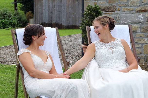 Два красиві нареченої на їх же секс весілля, сидів на дерев'яні шезлонгах, дивлячись один на одного і посміхаючись. Сільських, сільському церемонії в Йоркширський сільській місцевості  - Фото, зображення