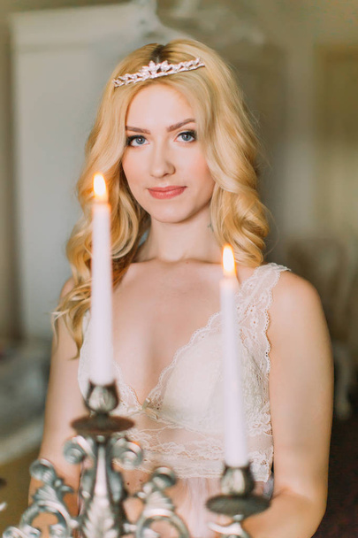 Portrait of sexy blonde bride in white underwear and diamond diadem on head  holding old candelabrum - Foto, Imagen