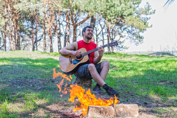 Young man playing acoustic guitar while sitting near campfire. - Foto, imagen