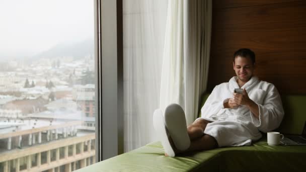 Young smiling handsome man wearing bathrobe messaging on a mobile phone and smiling. He is sitting near window with great city view with cup of coffee at luxury scyscraper hotel. - Footage, Video