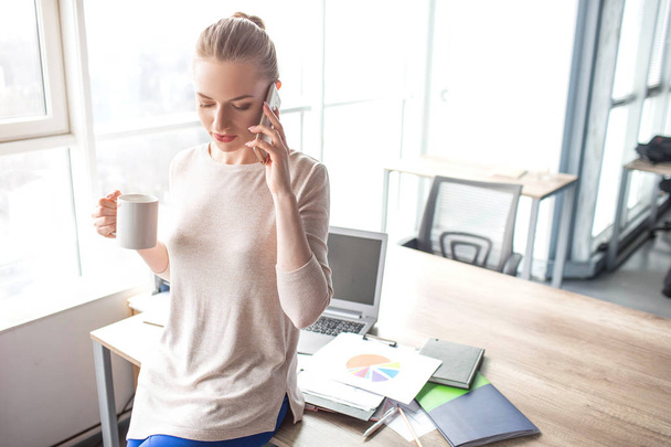 Business woman is standing at her office table and talking on the phone. She is holding a cup of tea in one hand and holding the phone with the other one. There are lots of notebooks on the table. - Foto, Imagem