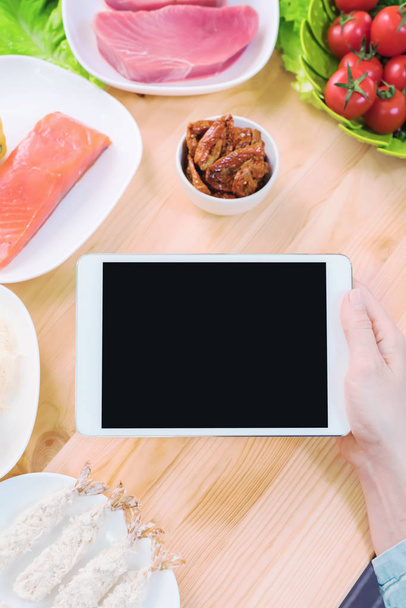 Close-up top view of female hands holding a tablet computer with a blank display surrounded by healthy food on a home kitchen table. Workpiece to insert the design of the mobile application into the - Foto, immagini