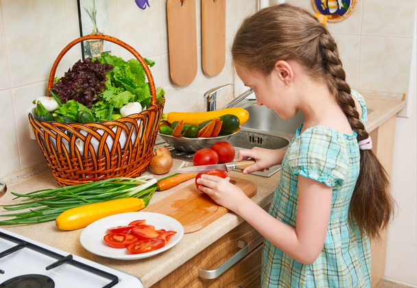 Παιδί κορίτσι μαγείρεμα στην κουζίνα στο σπίτι, για κοπή ντομάτες. Καλάθι με λαχανικά και φρέσκα φρούτα στο εσωτερικό της κουζίνας. Έννοια της υγιεινής διατροφής - Φωτογραφία, εικόνα