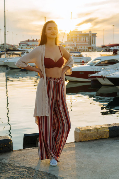 beautiful long-haired girl in trendy stylish pants standing in the port by the sea, in the evening sun, smiling - 写真・画像