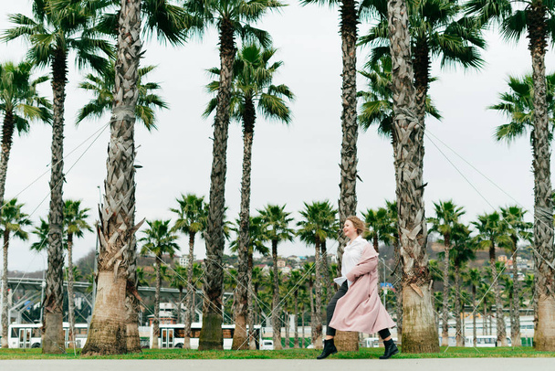 stylish girl in a pink coat walks in the park on a background of tall green palms, enjoying the long-awaited spring - Φωτογραφία, εικόνα