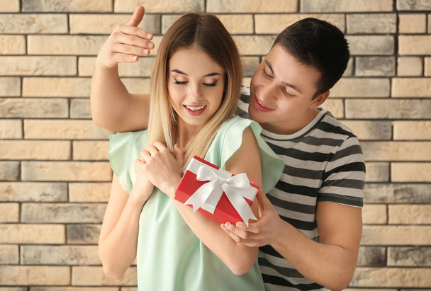 Young man giving present to his beloved girlfriend near brick wall - Фото, изображение