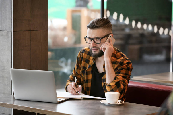 Young freelancer with mobile phone and laptop working in cafe - Photo, Image