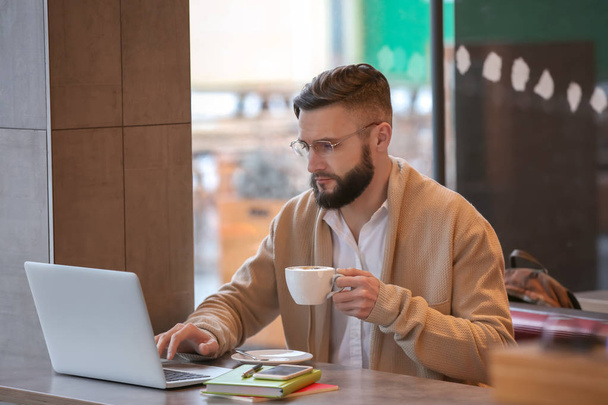 Молодой фрилансер с чашкой кофе и ноутбуком работает в кафе
 - Фото, изображение