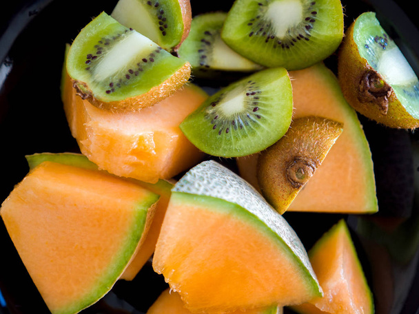 Close up food photograph of fresh ripe and mouth watering cantaloupe or muskmelon pieces with sliced kiwifruit served in a black bowl with vibrant orange and green colors. - Photo, Image