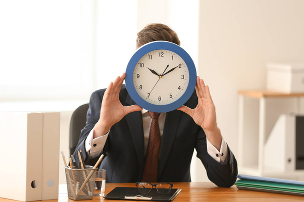 Man hiding face behind clock at table in office. Time management concept - Fotografie, Obrázek