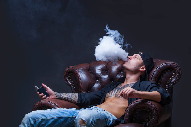 Vaper. The man with tattoos sits on a leather sofa smoke an electronic cigarette on the dark background. - Photo, Image