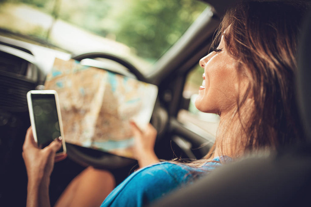 Rear view of young smiling woman in car looking at road map and using smartphone - Фото, изображение