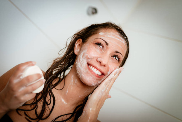 νέοι χαμογελαστό γυναίκα πρόσωπο πλύσιμο με σαπούνι στο μπάνιο - Φωτογραφία, εικόνα