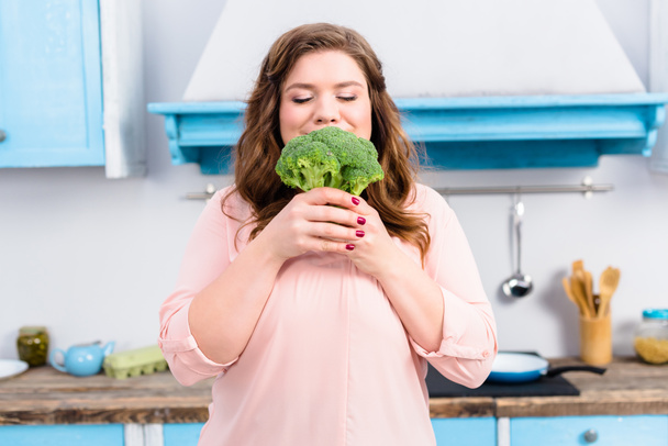 portrait of overweight young woman with fresh broccoli in hands in kitchen at home - Foto, imagen