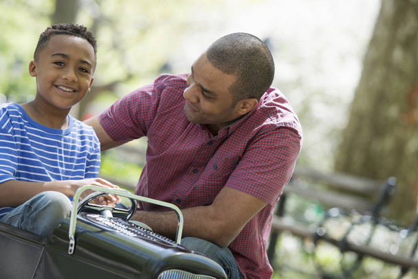 Father helping boy riding old fashioned toy peddle car in sunny park. - Photo, Image