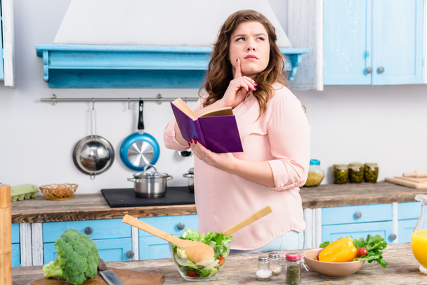 zamyślony kobieta z książka kucharska stojący w kuchni w domu z nadwagą - Zdjęcie, obraz