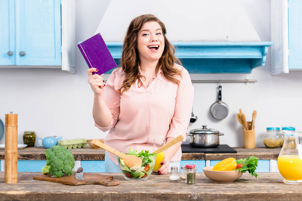Übergewichtige junge Frau mit Kochbuch steht zu Hause in Küche - Foto, Bild