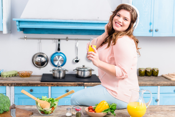uśmiechający się nadwagę Kobieta w słuchawkach z szklankę soku w parze w kuchni - Zdjęcie, obraz