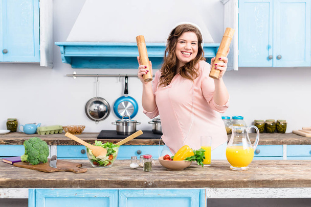 cheerful overweight woman in headphones with wooden salt and pepper grinders in hands in kitchen - Foto, immagini