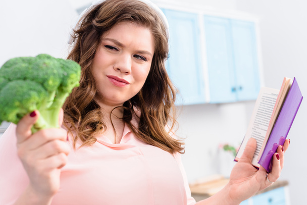 Übergewichtige Frau mit Kopfhörer und Kochbuch blickt in der Küche auf frischen Brokkoli in der Hand - Foto, Bild