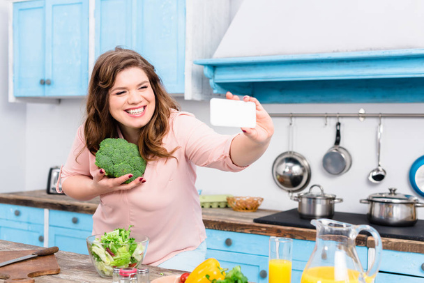 mulher sorrindo sobrepeso com brócolis na mão tomando selfie no smartphone na cozinha em casa
 - Foto, Imagem