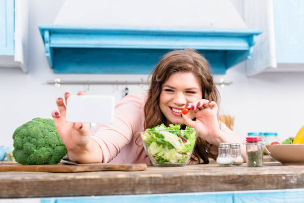 Übergewichtige lächelnde Frau mit Kirschtomate in der Hand macht Selfie auf Smartphone in der heimischen Küche - Foto, Bild