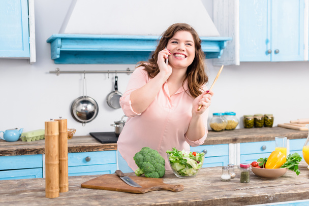 mujer joven con sobrepeso con cuchara de madera en la mano hablando en el teléfono inteligente mientras está de pie en la mesa con verduras frescas en la cocina en casa
 - Foto, Imagen