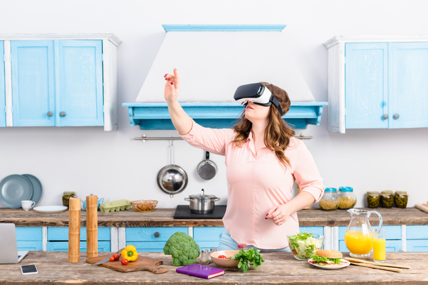 overweight woman in virtual reality headset standing at table with fresh vegetables in kitchen at home - 写真・画像