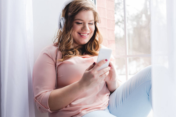 jovem mulher sorridente em fones de ouvido usando smartphone em casa
 - Foto, Imagem