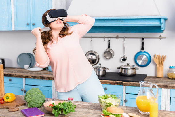 young woman with knife in virtual reality headset standing at table with fresh vegetables in kitchen at home - Photo, image