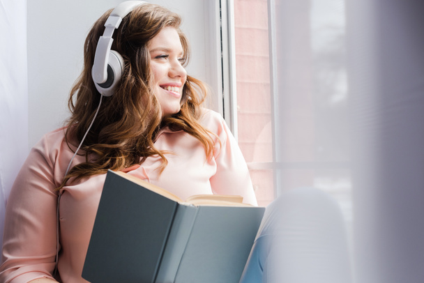 mujer sonriente en auriculares con libro sentado en el alféizar de la ventana en casa
 - Foto, Imagen