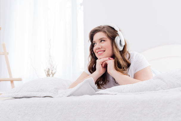usmívající se žena v pyžama poslech hudby ve sluchátkách na posteli doma - Fotografie, Obrázek