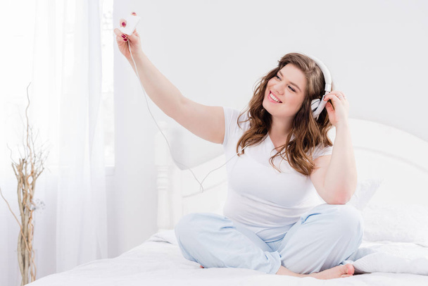 young smiling woman in headphones taking selfie on smartphone on bed at home - Fotó, kép
