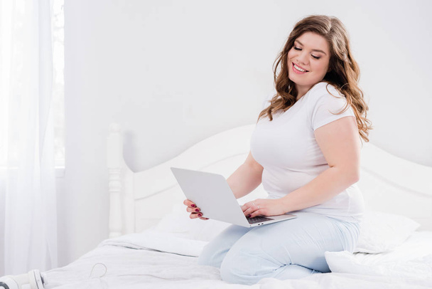 usmívající se žena v pyžama pomocí přenosného počítače na posteli doma - Fotografie, Obrázek