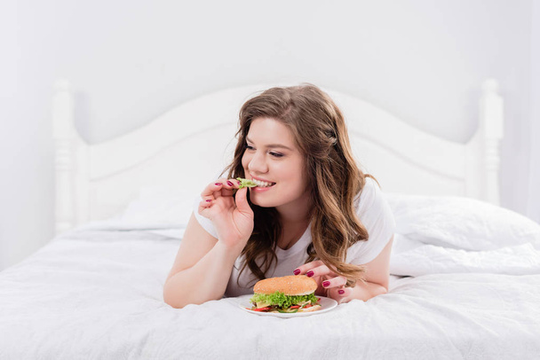 overweight woman in pajama eating burger on bed at home - Photo, Image