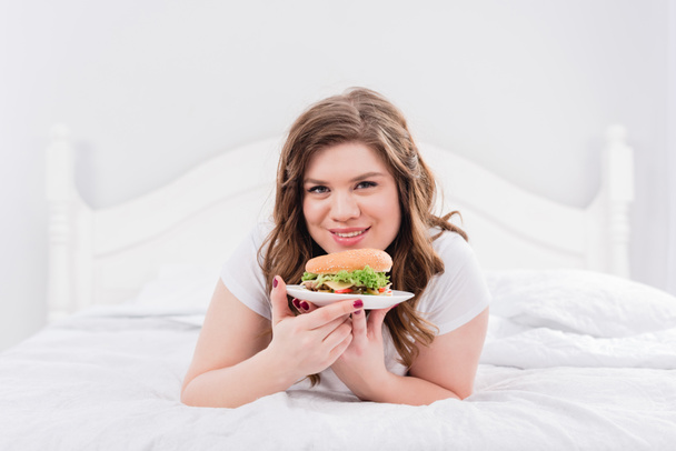 portrait of overweight smiling woman in pajama with burger on bed at home - Foto, imagen