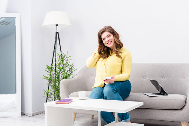 young smiling woman with smartphone sitting on sofa with laptop at home - Fotoğraf, Görsel
