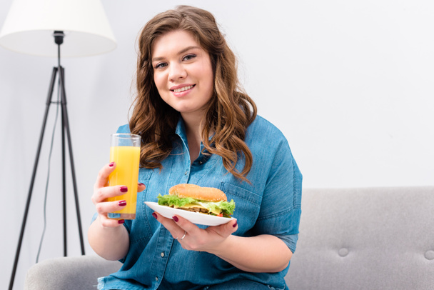 overweight smiling woman with glass of juice and burger on plate in hands at home - Fotografie, Obrázek