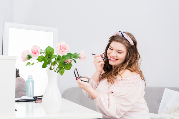 pretty smiling young woman applying lip gloss while doing makeup at home - Foto, Imagen