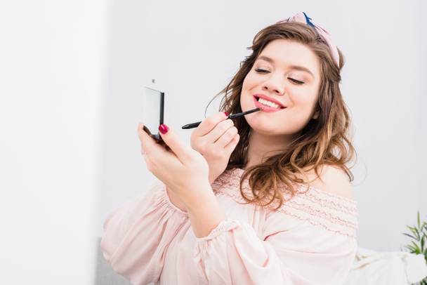 portrait of pretty smiling young woman applying lip gloss while doing makeup at home - Photo, Image