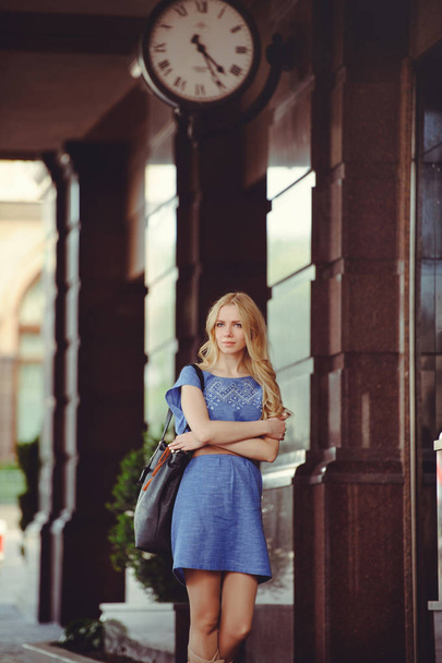 schöne blonde Frau steht unter dem Haus mit einer großen Uhr in einem blauen Kleid und Stiefeln gekleidet, bescheiden und schüchtern - Foto, Bild
